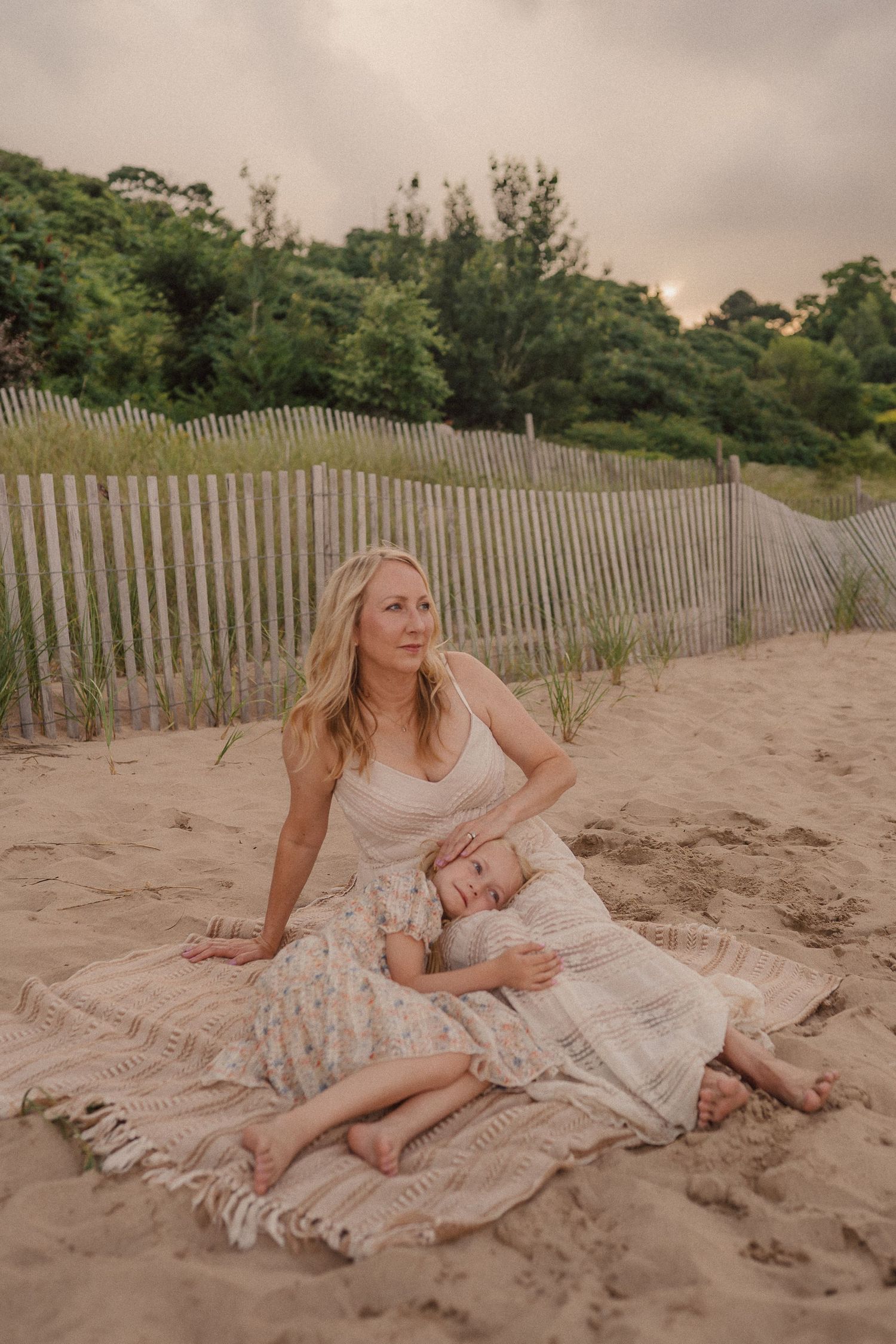 Woman in flowing dress poses on sandy beach against weathered wooden fence at sunset.