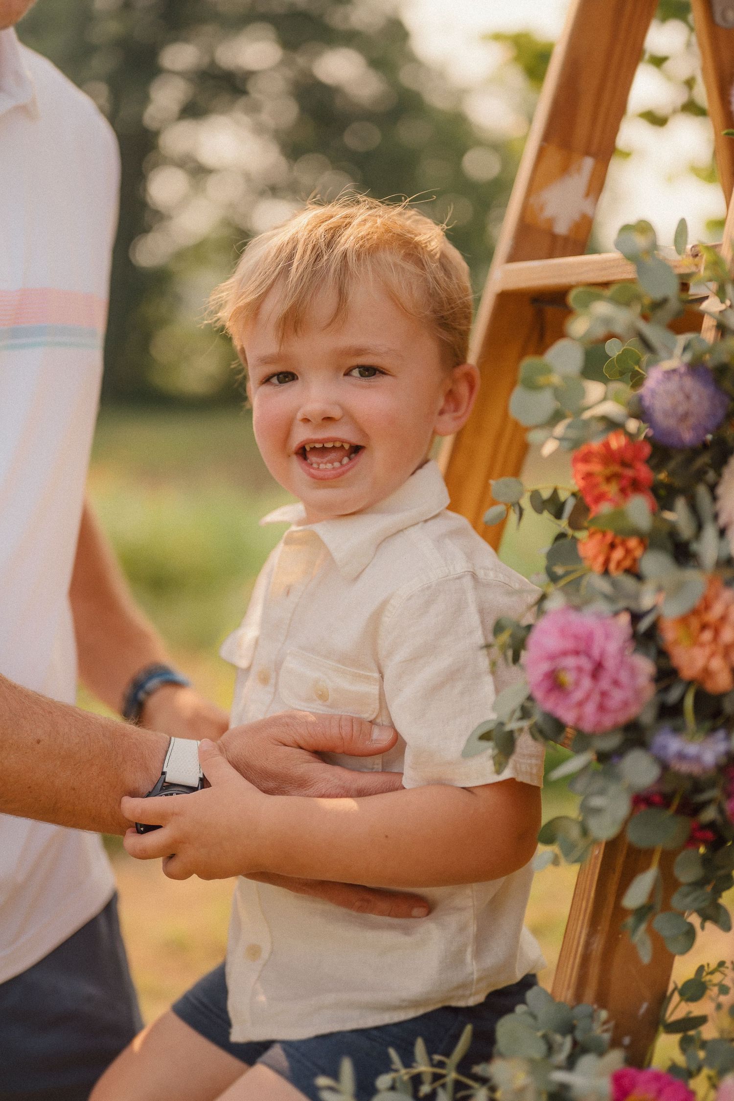 A happy toddler in a white shirt laughs during an outdoor event decorated with colorful floral arrangements.