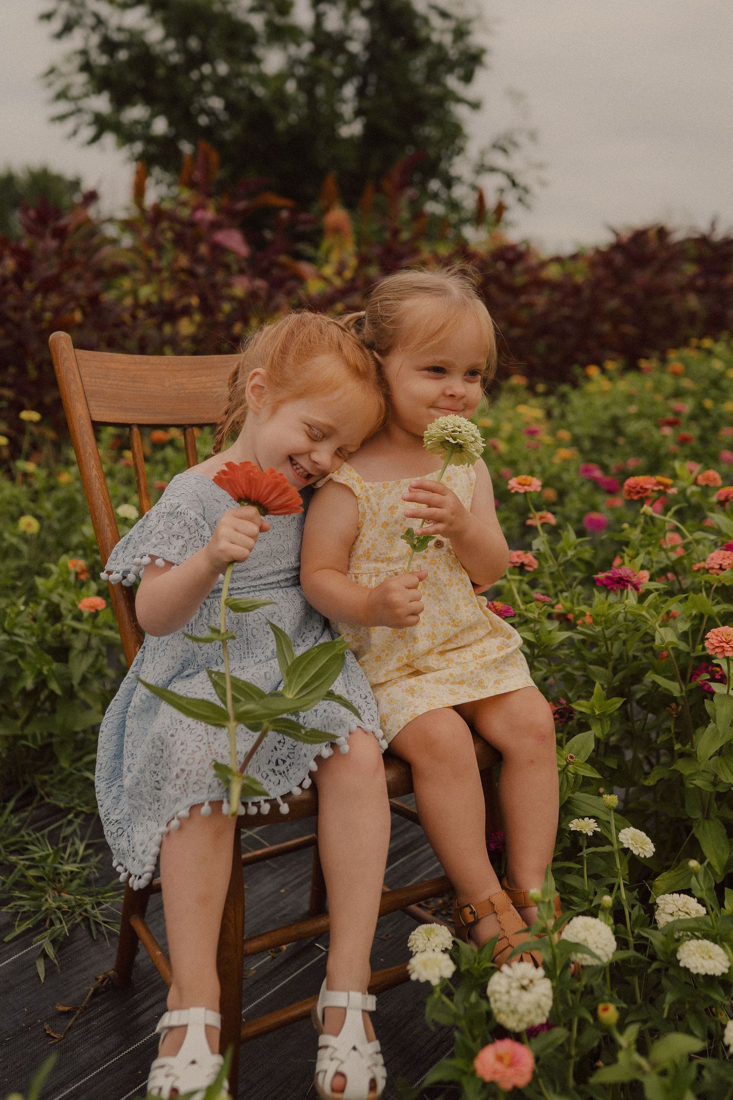 Two small children share a moment on a bench in a blooming flower garden.