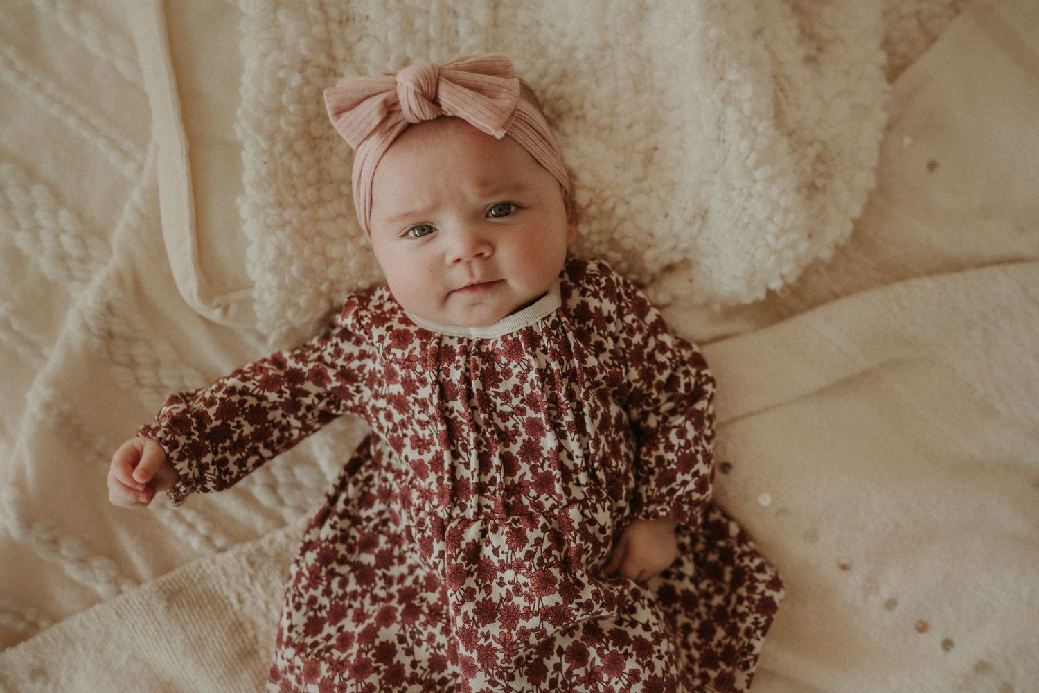 Baby wearing a floral dress and pink bow headband lying on white bedding.