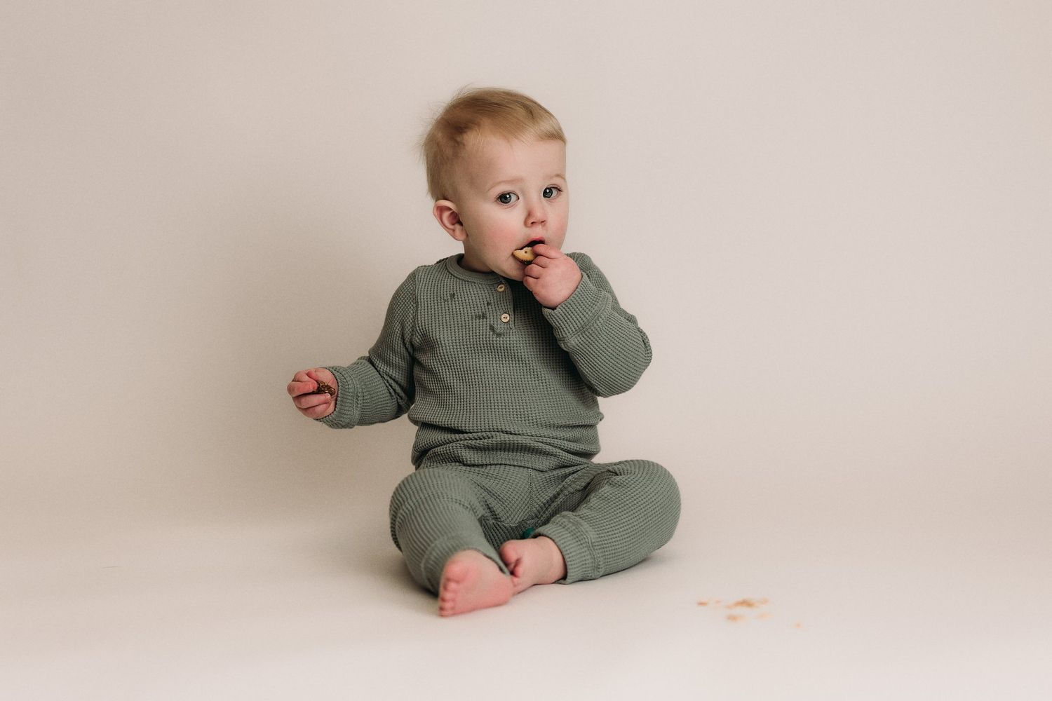Baby in sage green ribbed outfit sits on cream backdrop with finger in mouth.
