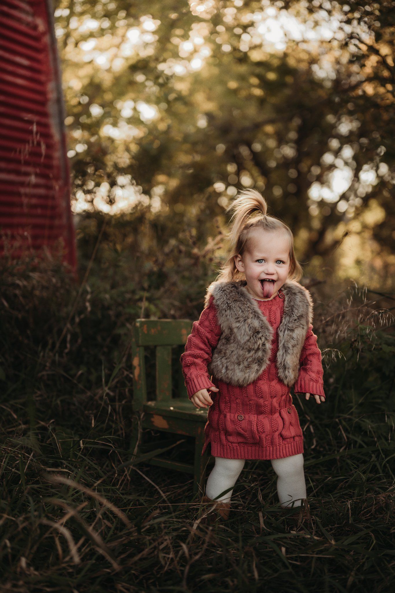 Child wearing red sweater and fur vest laughing outdoors near rustic barn.