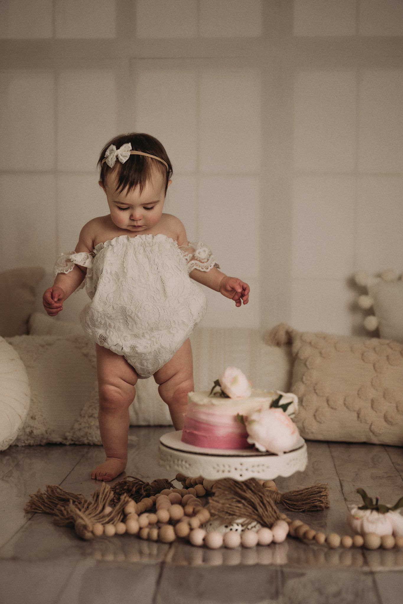 Baby in lace outfit dancing near pink cake with wooden bead garland decorations.