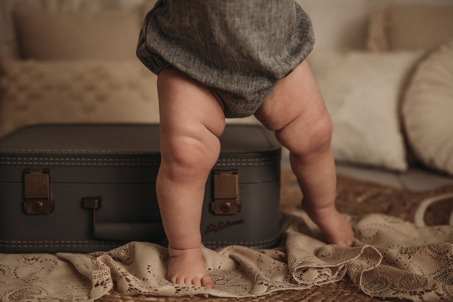 Small chubby legs in a gray romper standing on a vintage suitcase with lace fabric underneath.