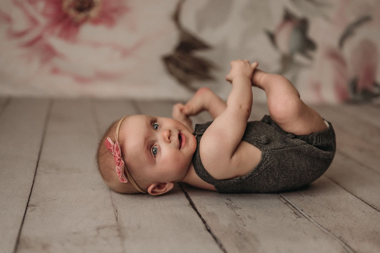 Baby in gray outfit lying on wooden floor beside floral backdrop.