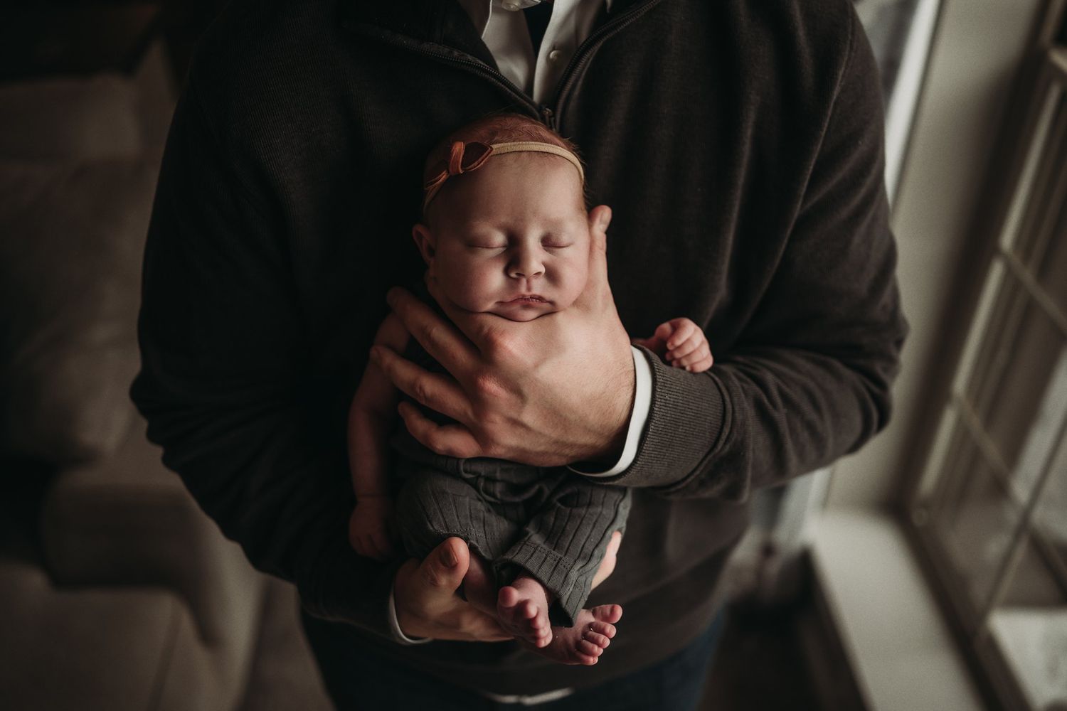 Close-up portrait of tiny newborn baby being held in brown sweater-clad arms.
