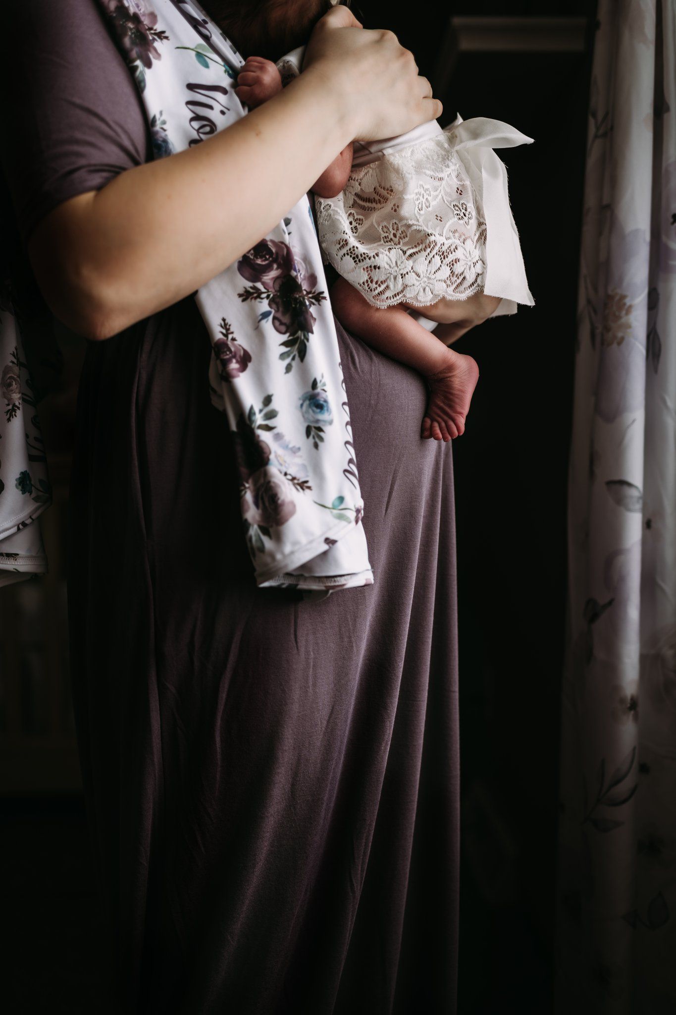 Person in floral dress holding swaddled baby in dim lighting.