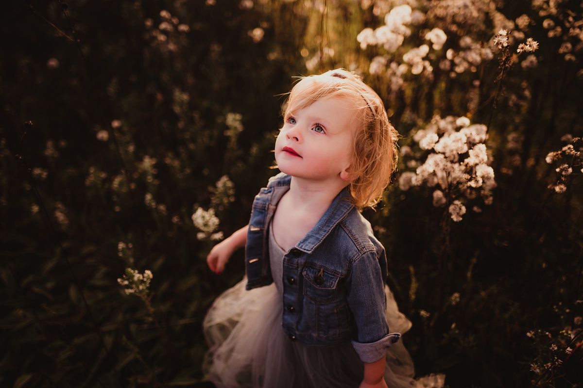 Child in denim jacket stands among blooming flowers in golden light.