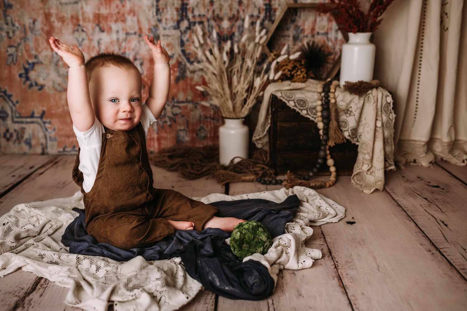 Sitting on wooden floor in brown overalls with arms raised against brick wall backdrop.