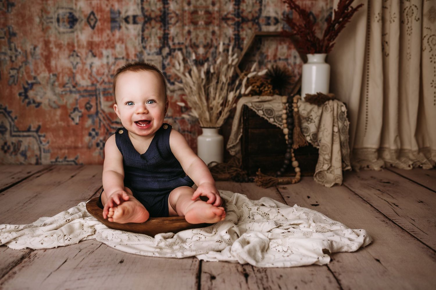 Joyful baby in navy romper sits in wooden bowl with vintage-style photography setup.