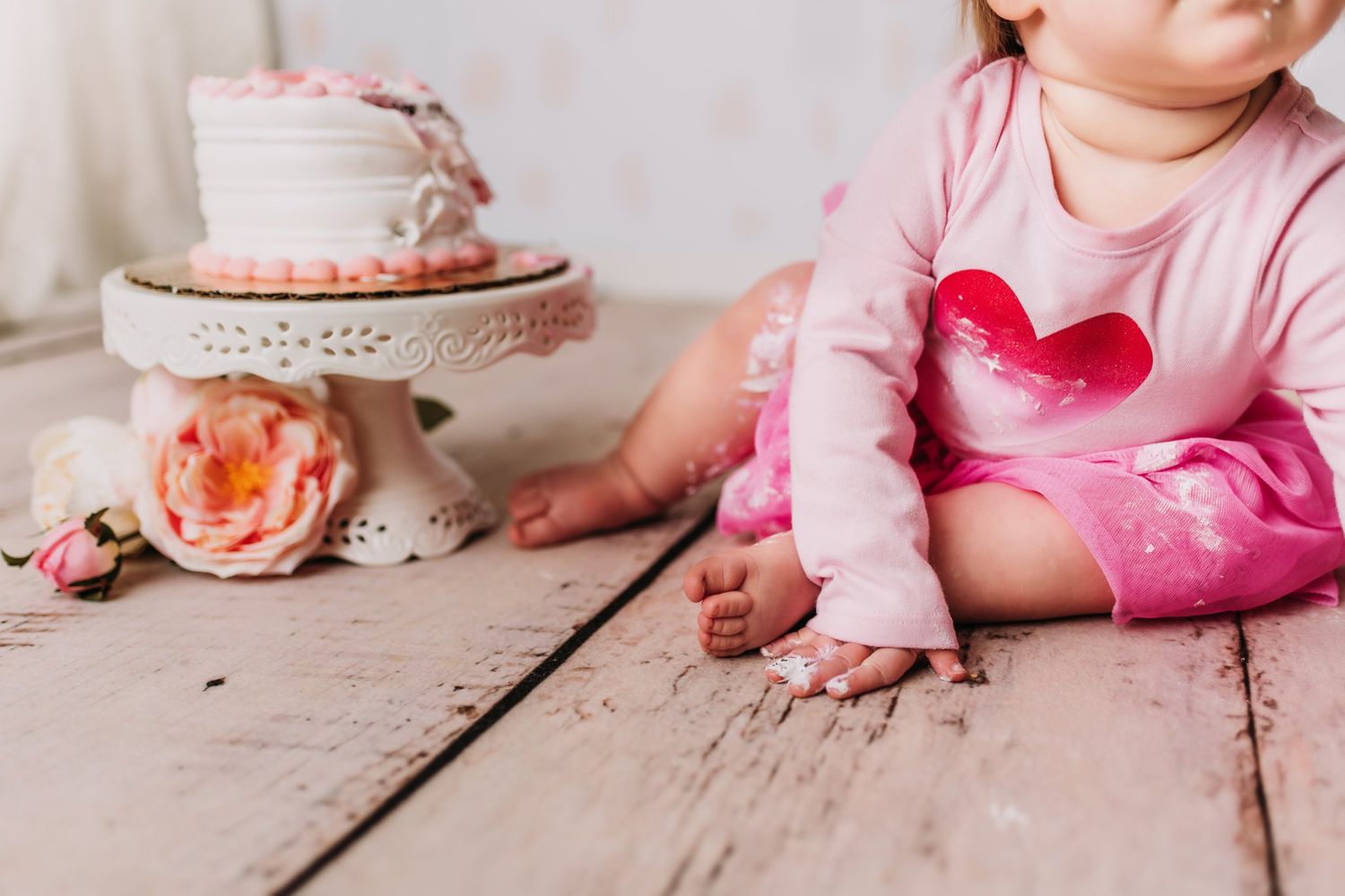 Close-up of pink cake on stand with peach roses and pink clothing details on wooden floor.
