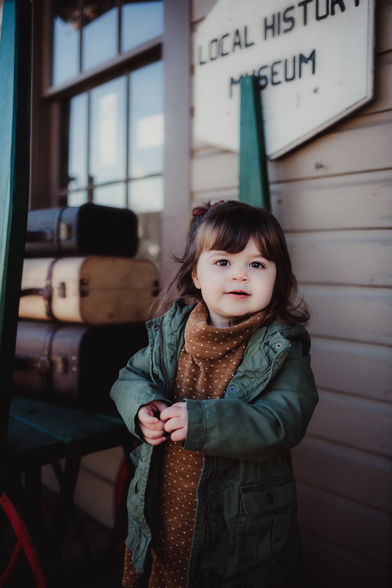 A toddler in a green coat and brown dress poses in front of a local history museum sign.