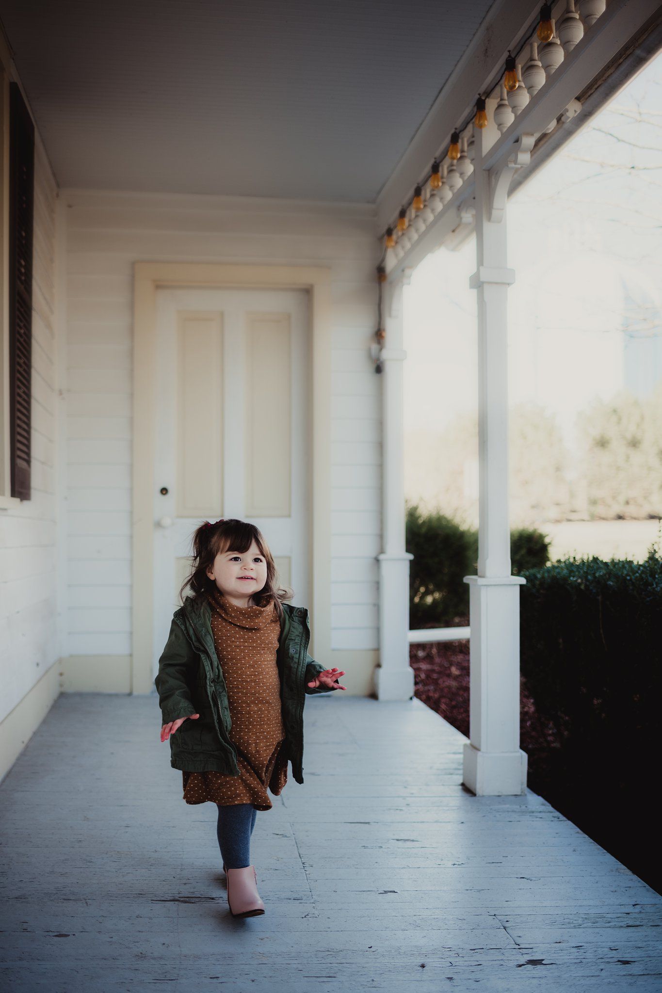 A small child in a green jacket stands on a white porch near classic architectural columns.