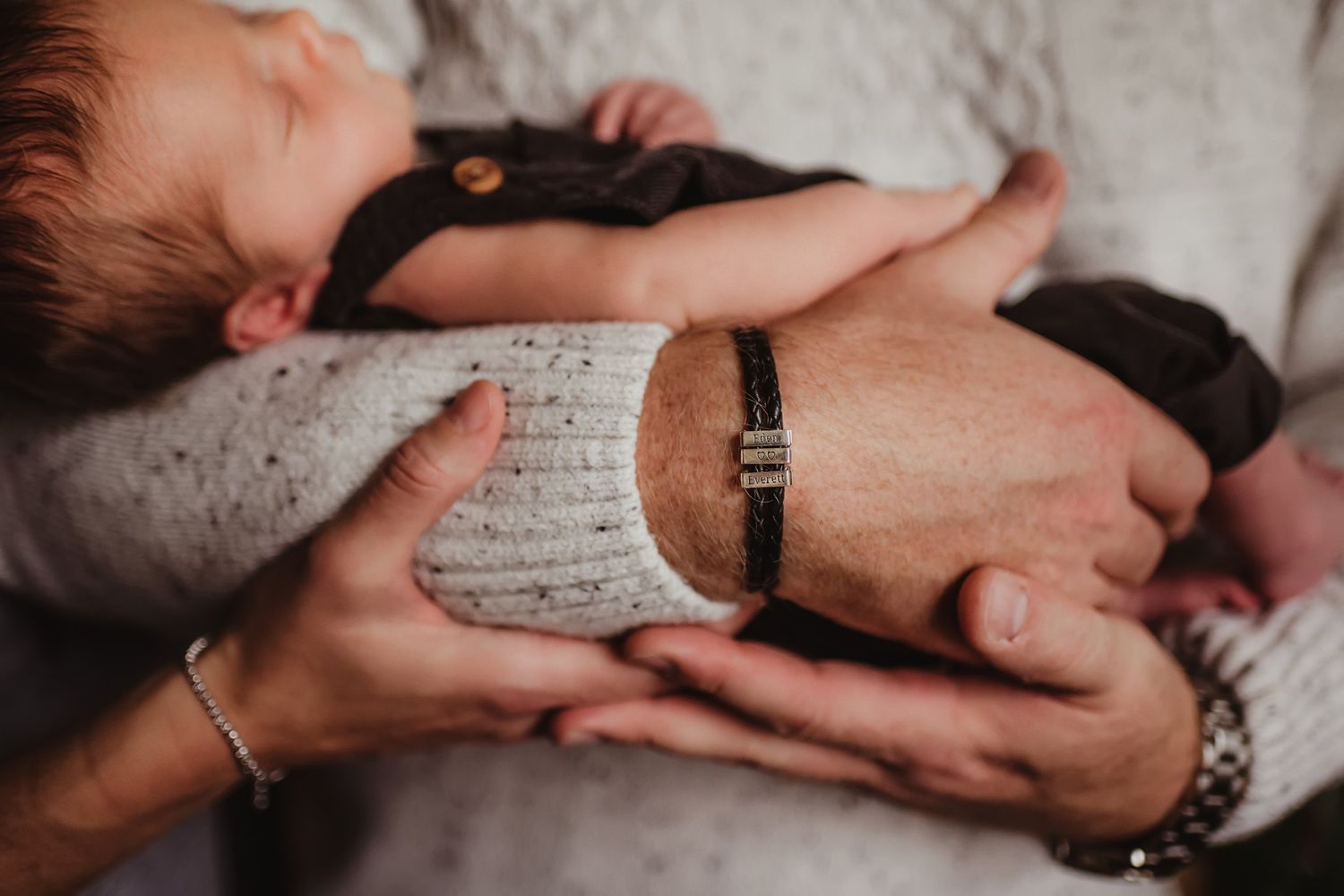 Close-up detail of hands tenderly holding a sleeping newborn baby wearing dark clothing.