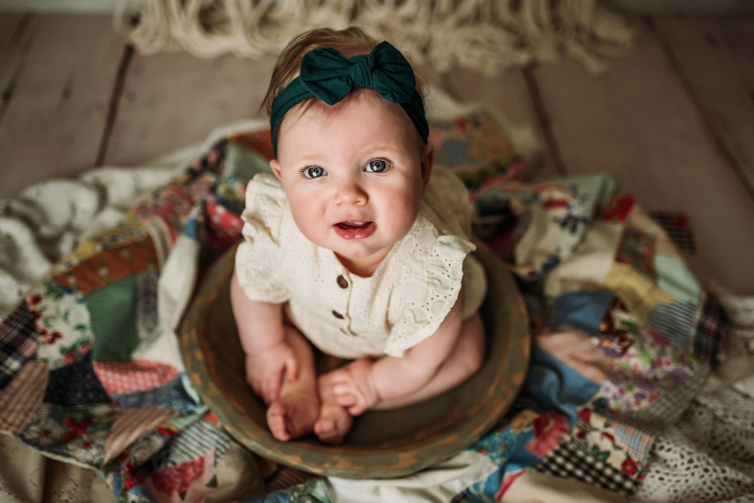 Baby wearing a white top sits in a wooden bowl surrounded by colorful quilted blankets.
