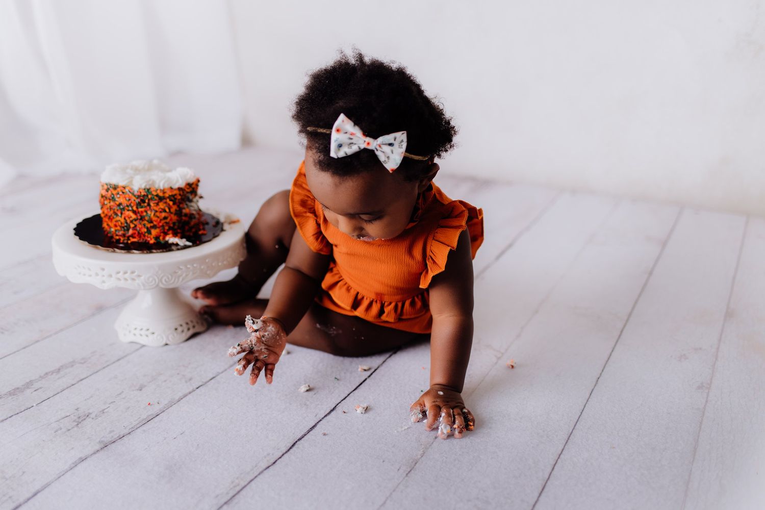 A small child in an orange dress crawls near a cake during a cake smash photo session.