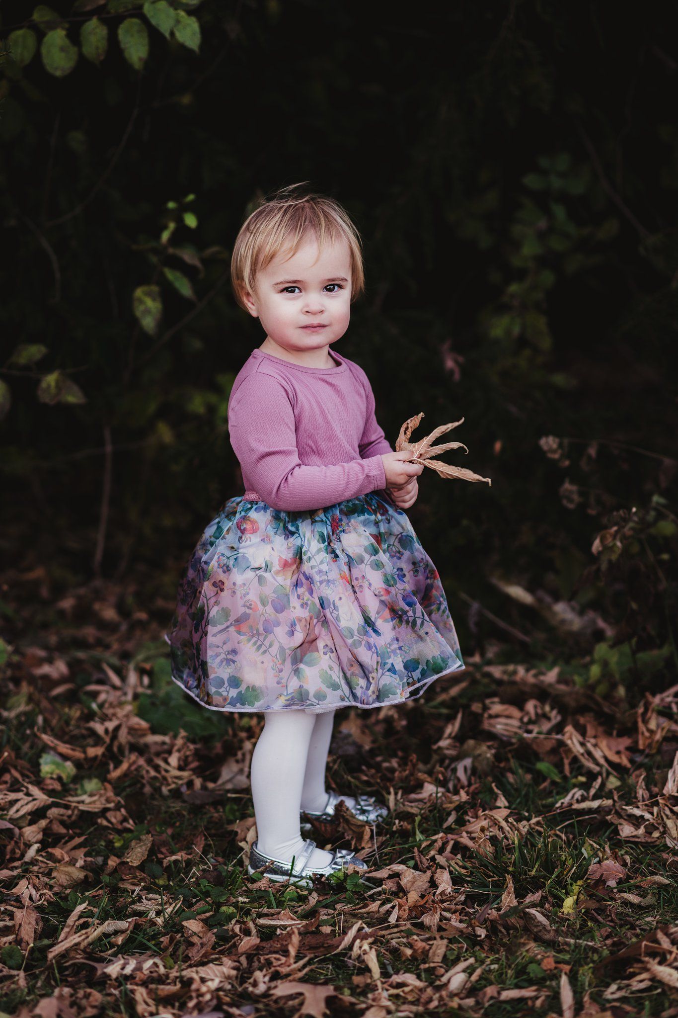 Small child in pink sweater and floral dress stands among fallen leaves.