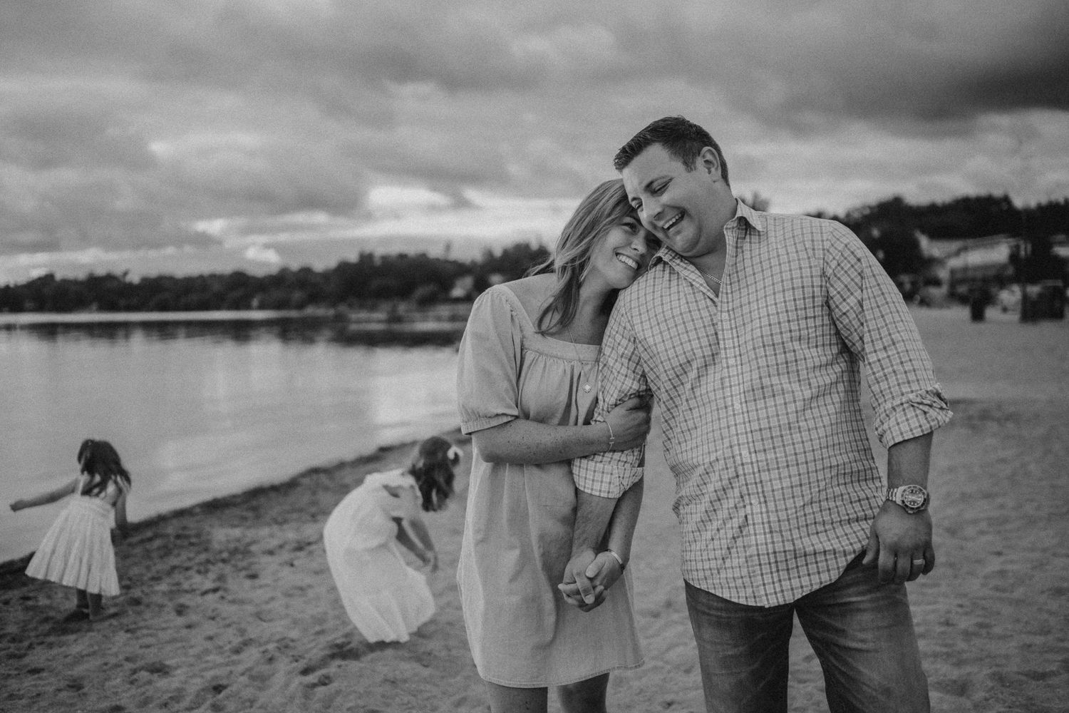 A couple shares a laugh on the beach while children play in the background.