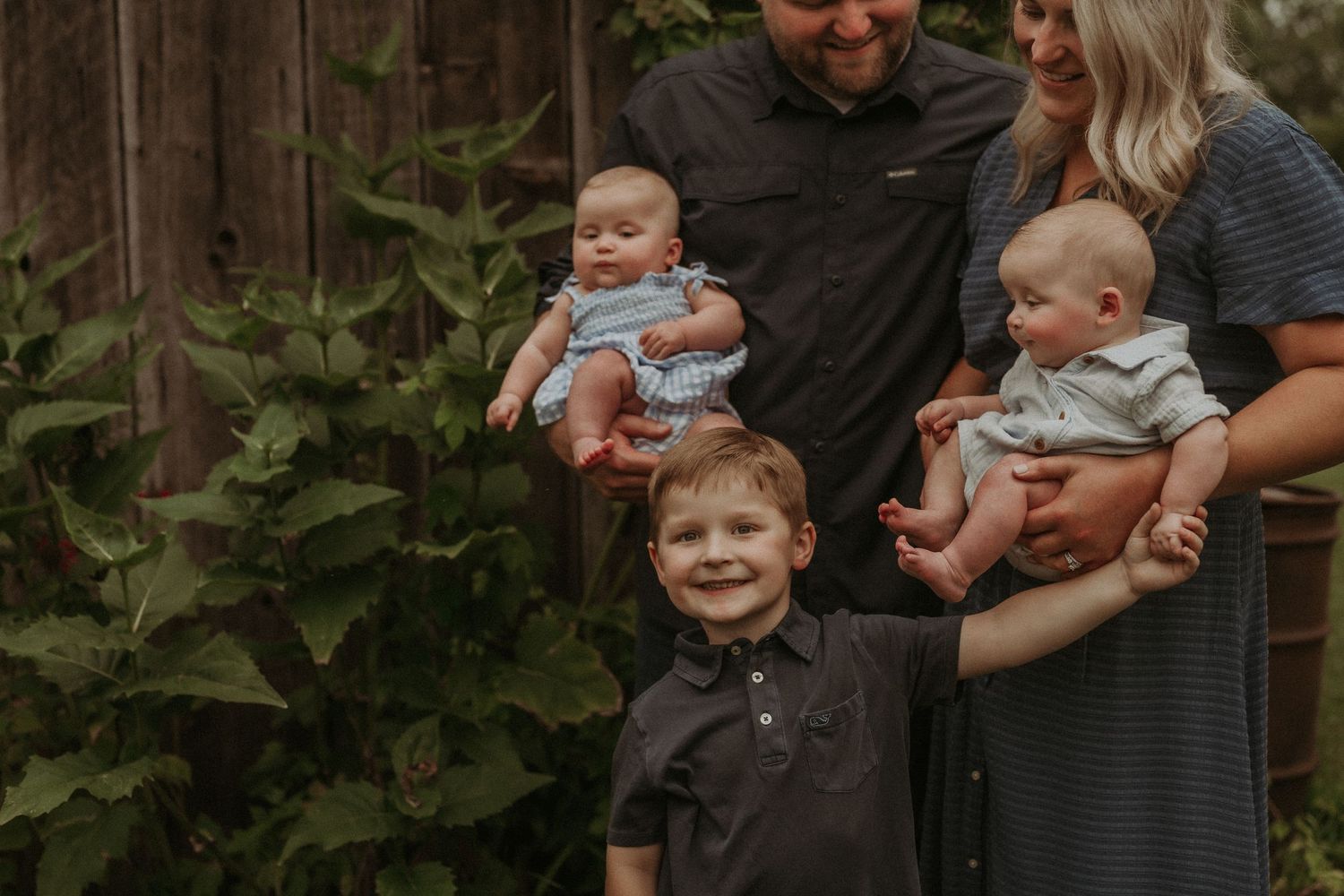 A family portrait in front of greenery shows parents holding twin babies while an older child stands in front.