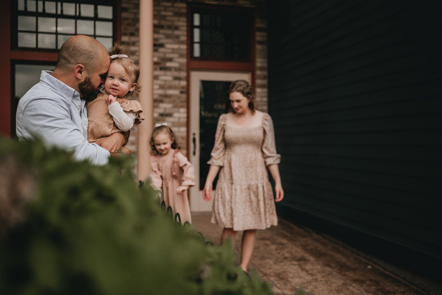 A family dressed in coordinating pink and neutral tones pose together outside a brick building.