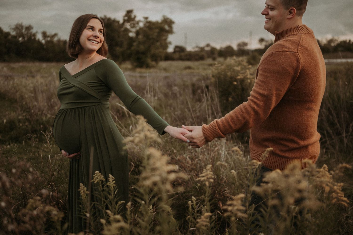Joyful maternity moment captured as couple holds hands in rustic field setting.