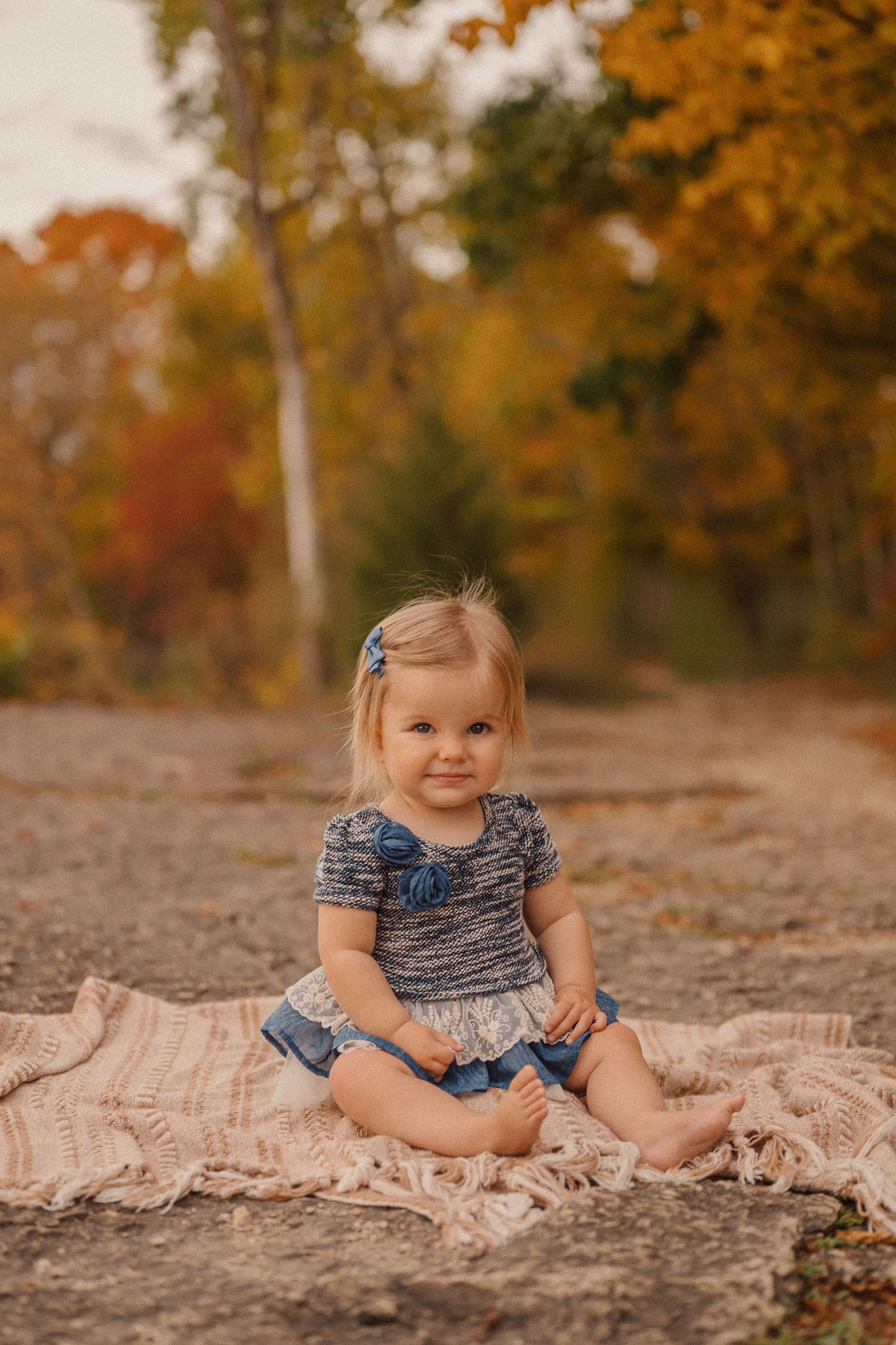 A baby in a striped dress sits on a beige blanket during an autumn outdoor photoshoot.