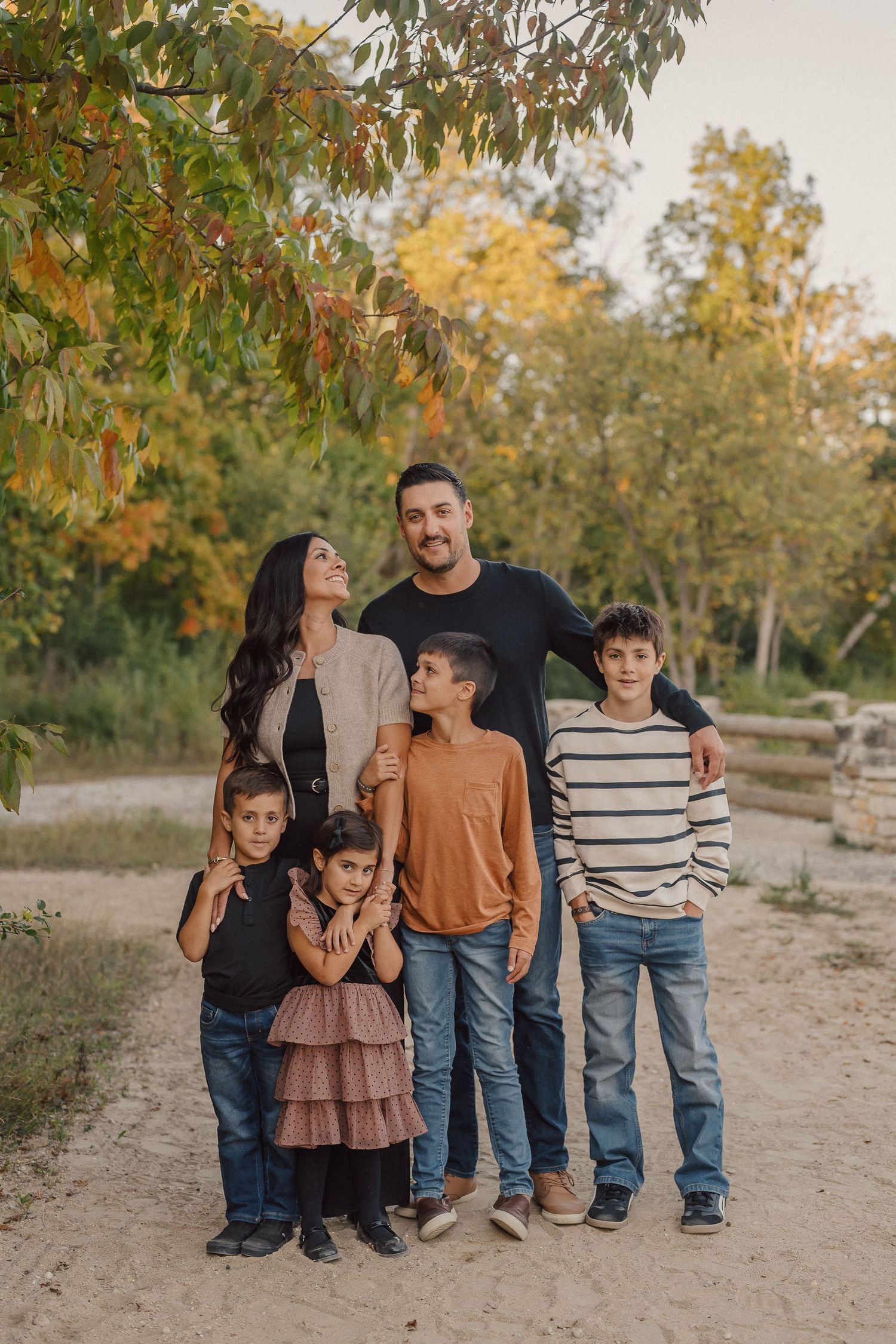 A family stands together on a dirt path surrounded by autumn trees during an outdoor photoshoot.