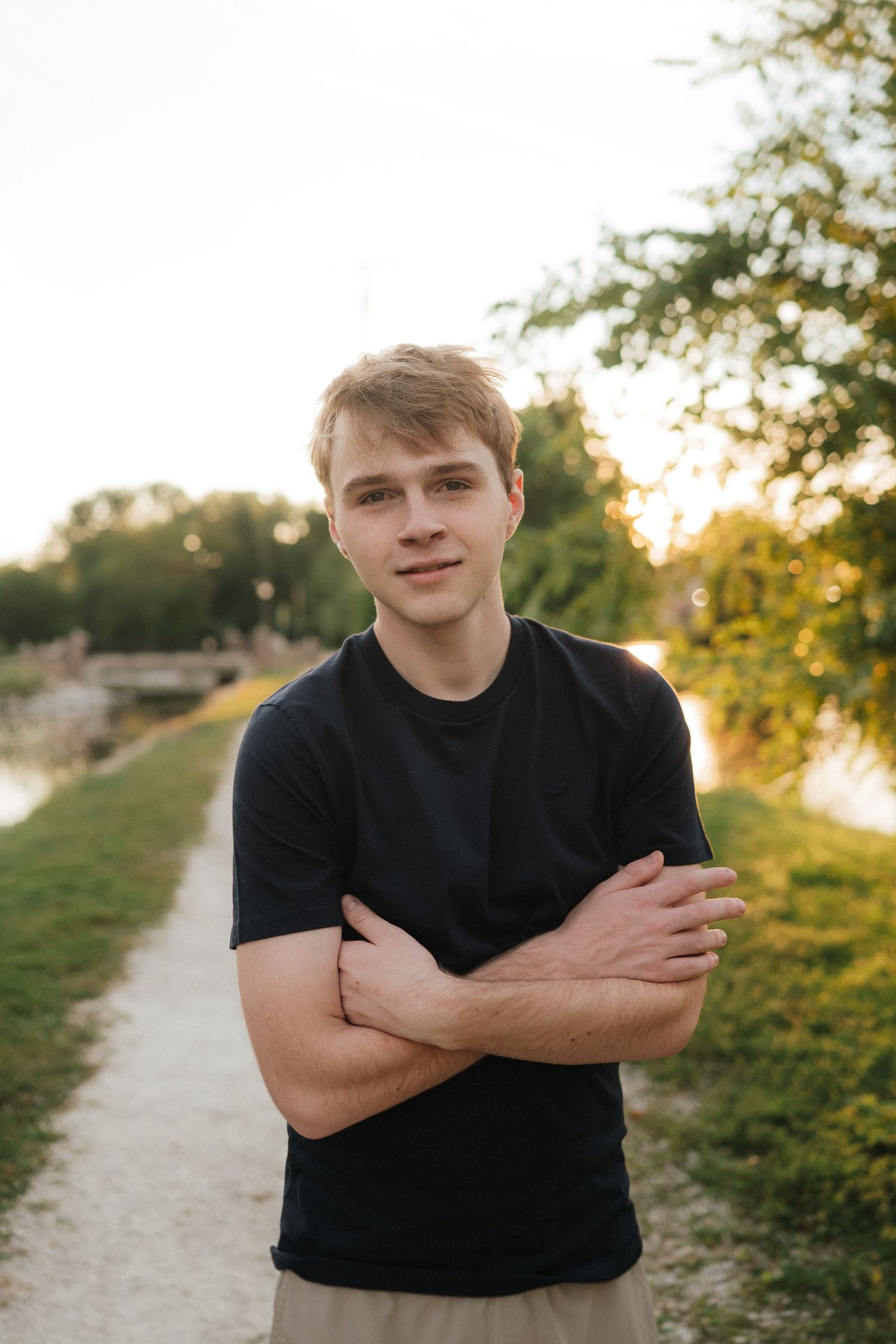 A person in a black t-shirt stands with arms crossed on a path at sunset.