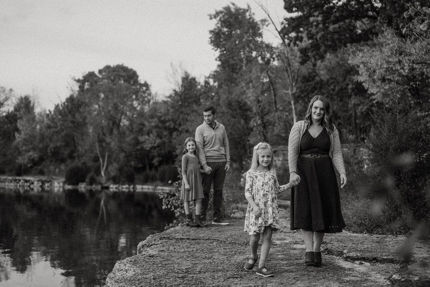 A family stands together on a rocky path beside a serene lake in black and white.
