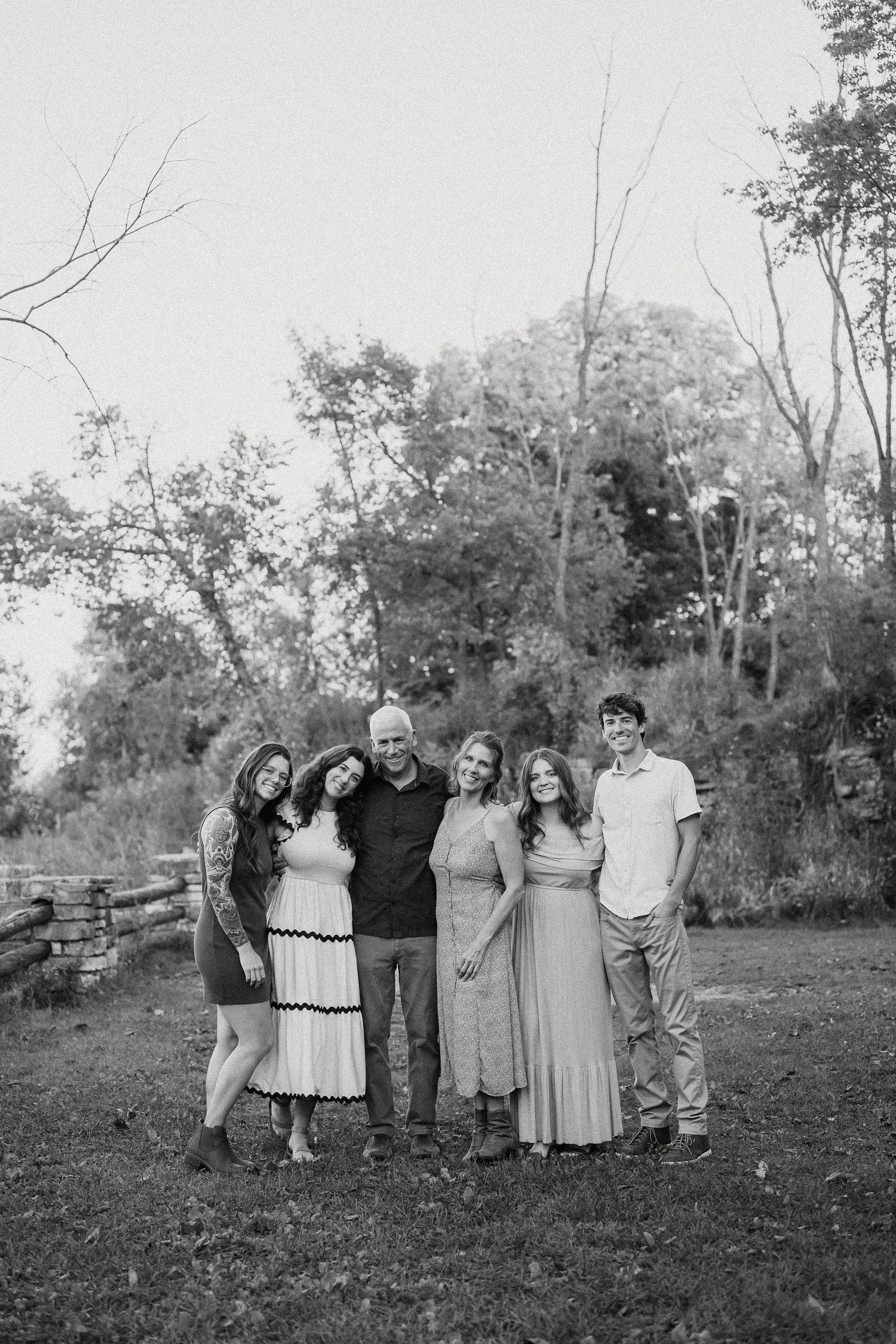A black and white outdoor family portrait showing a group of six people standing together on grass with trees in background.
