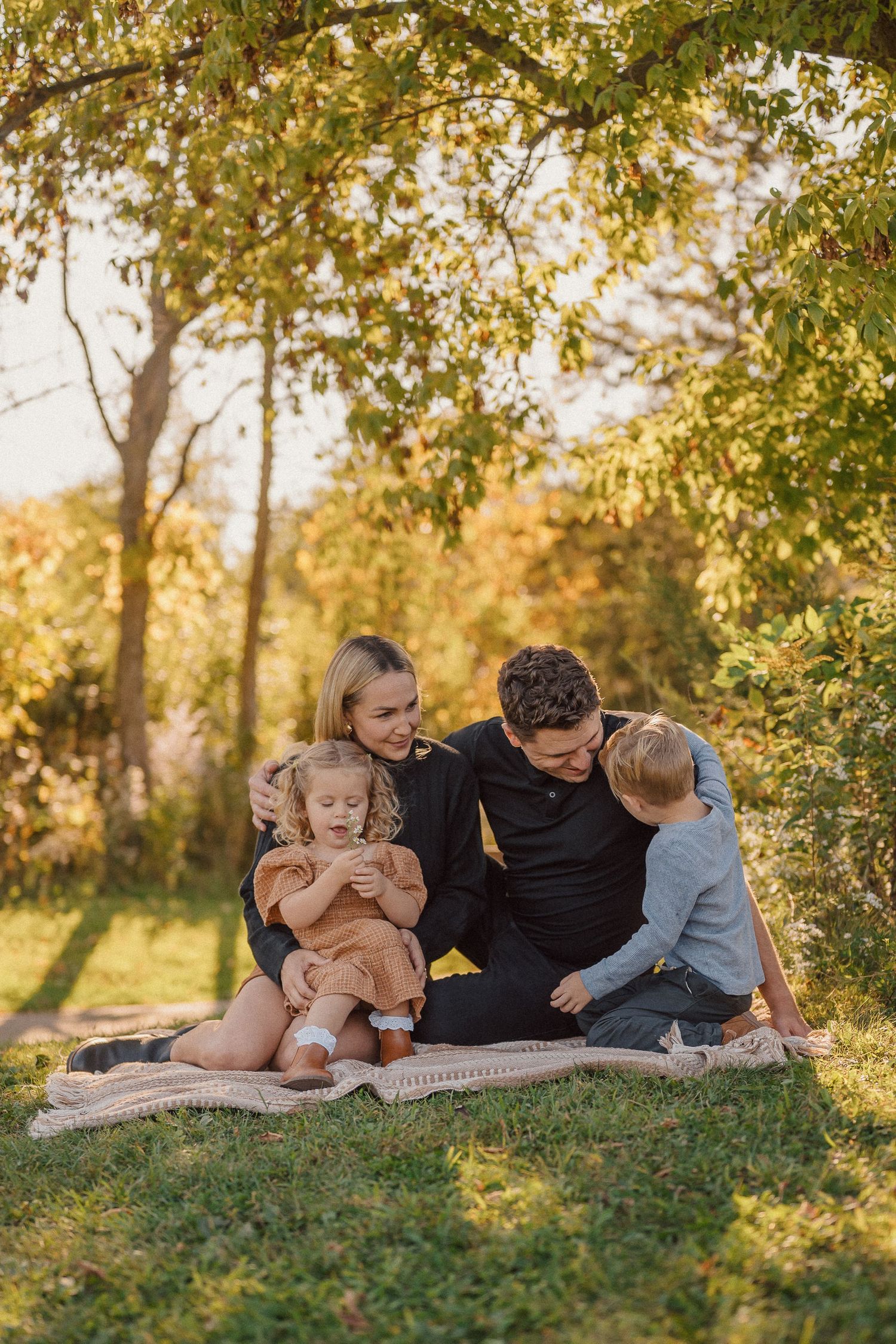 Family relaxes together on grassy hillside under trees during golden hour.
