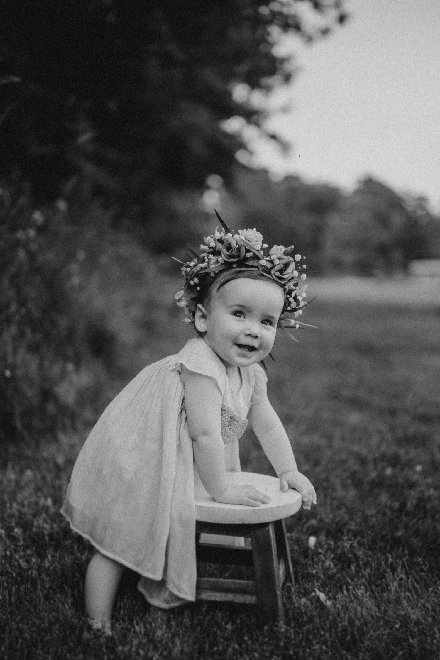 Black and white photo of small child in dress leaning on wooden stool wearing floral crown.