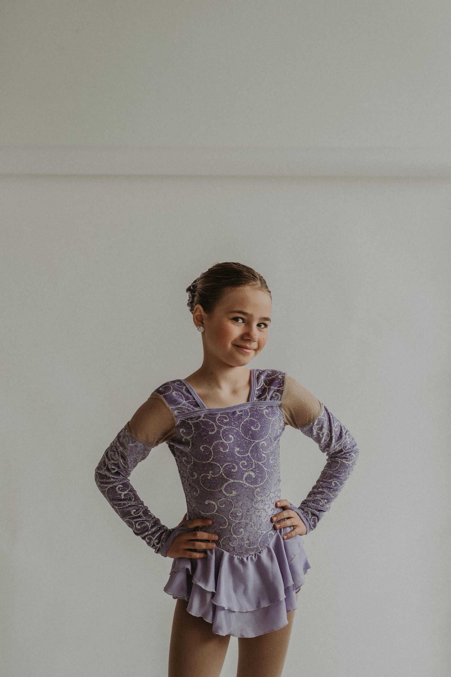 Figure skater poses in a purple performance dress with swirled pattern and layered skirt against white backdrop.