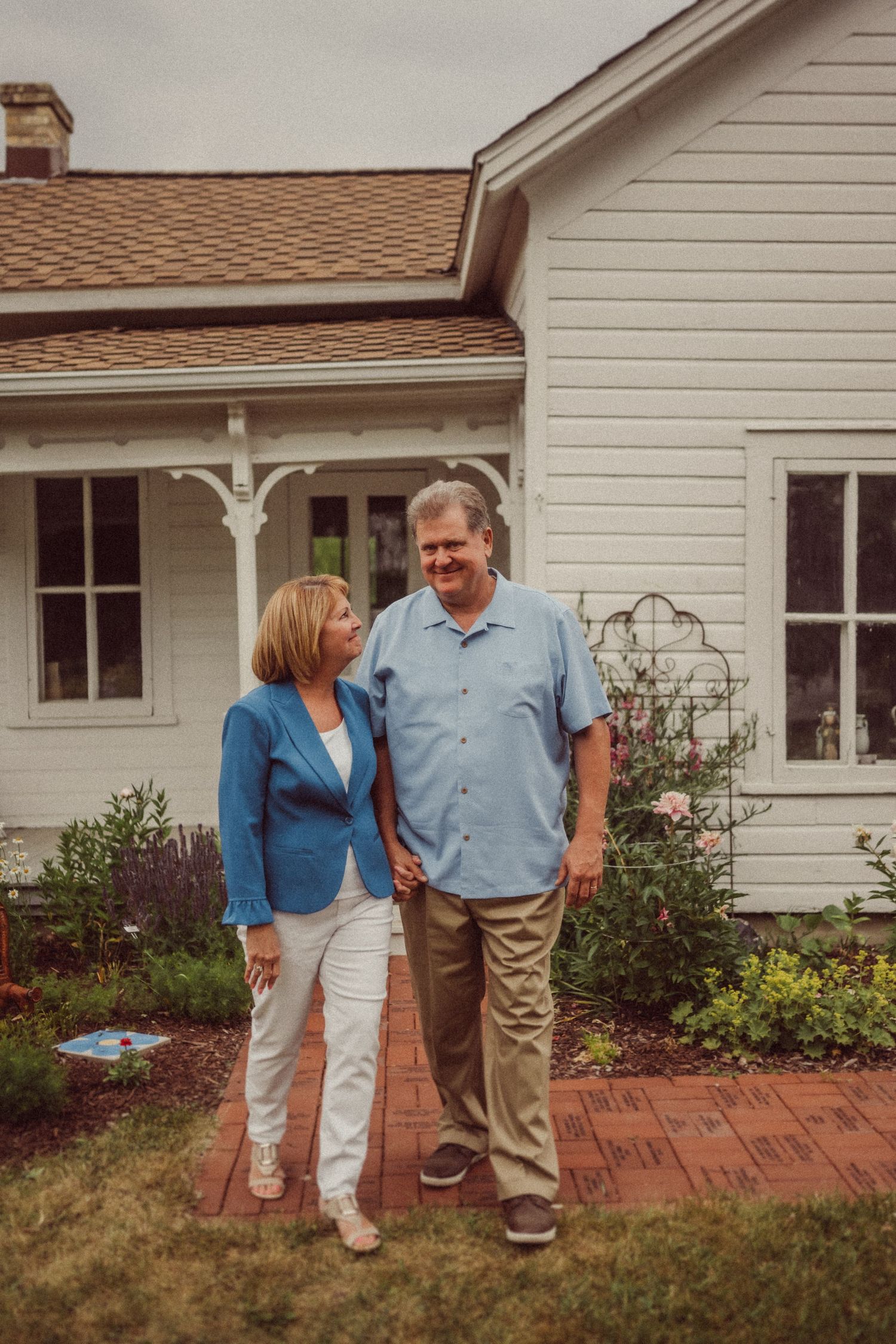 A couple stands together smiling outside their white farmhouse with Victorian-style porch trim.