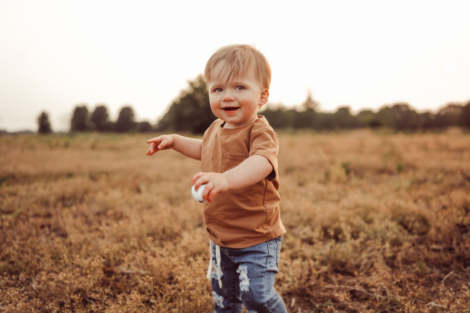 A toddler in a brown shirt and jeans plays joyfully in a rustic field setting.