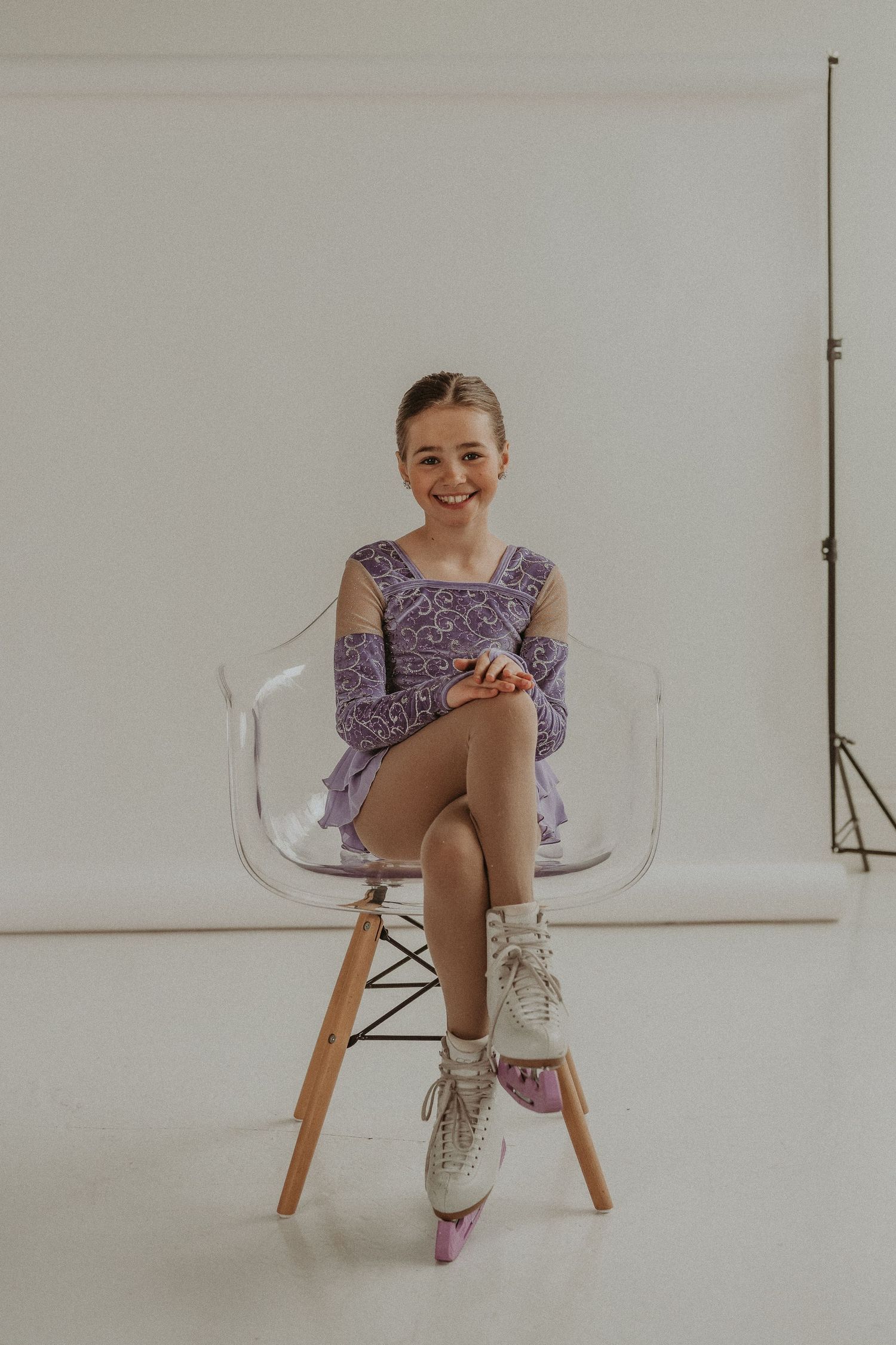 Ice skater in purple costume sits elegantly on clear modern chair in studio setting.