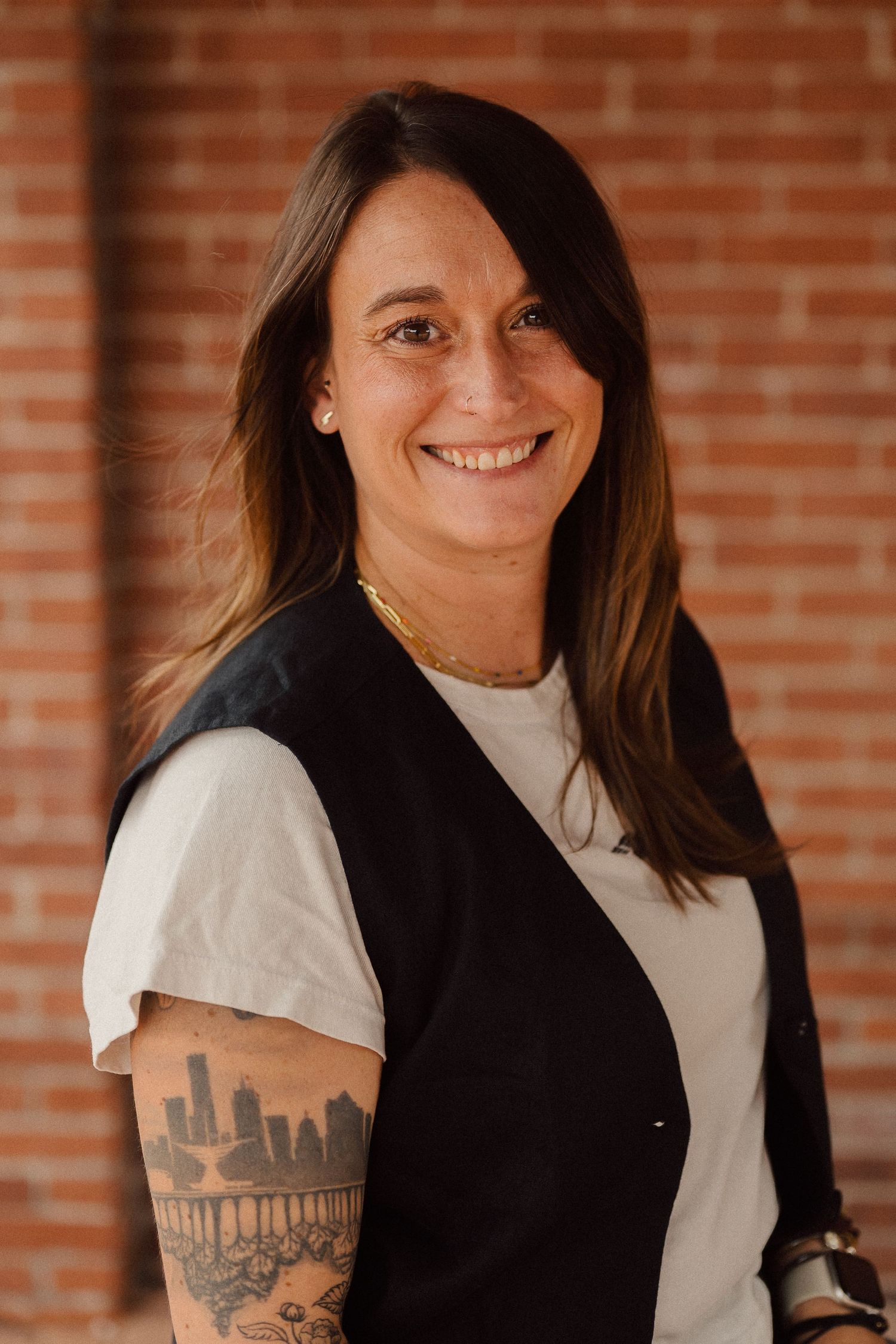 Professional portrait against brick wall showing a person in black vest and white shirt with visible arm tattoo.