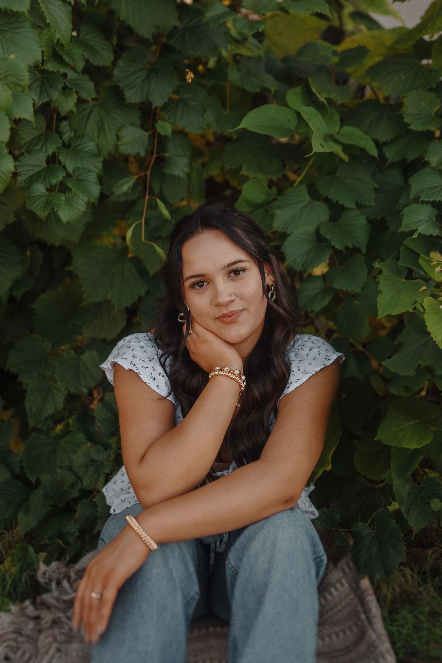 A person in a white lace top and jeans sits against a lush green grapevine backdrop during golden hour.