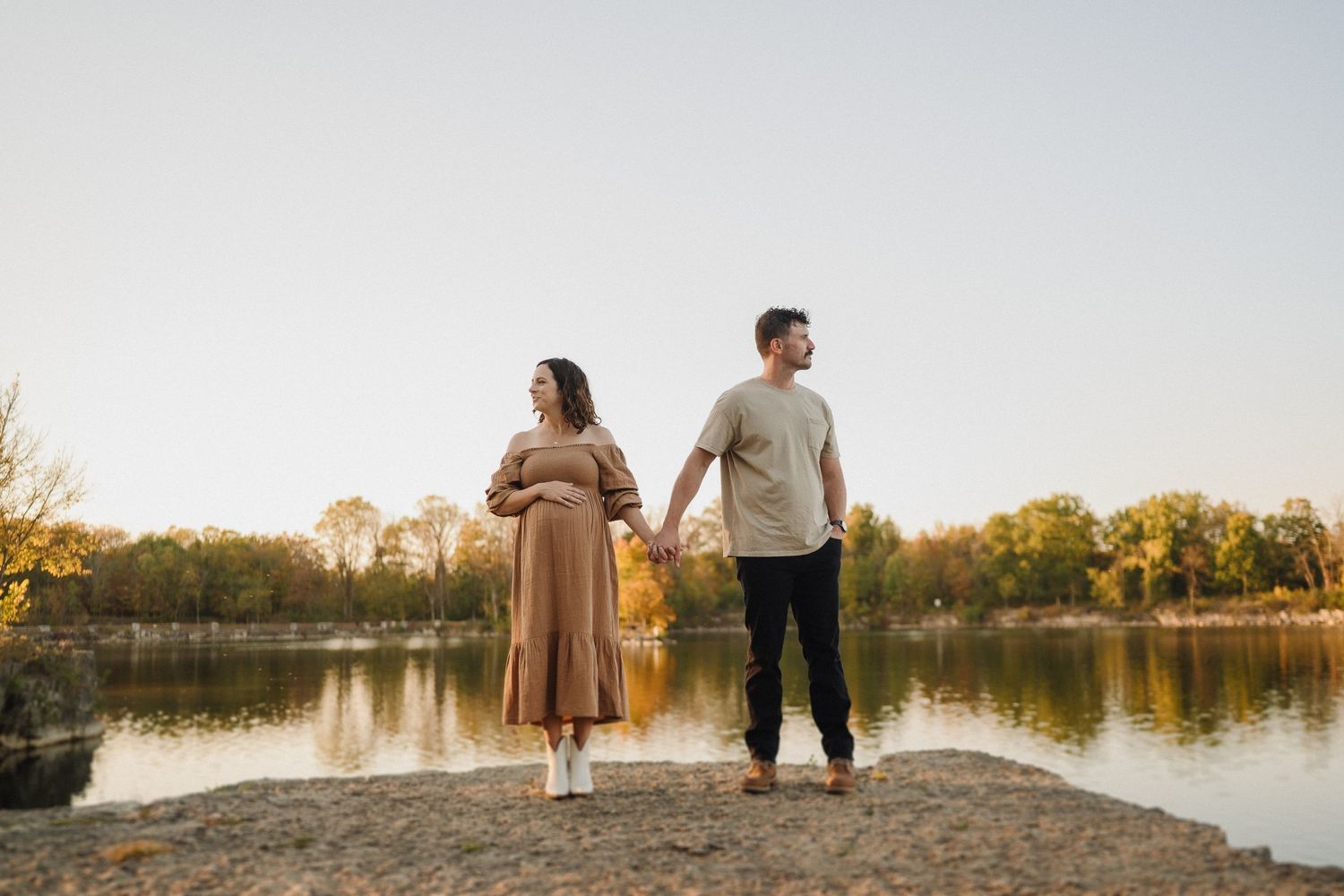 Expectant couple stands on rocky shore overlooking serene lake at sunset.