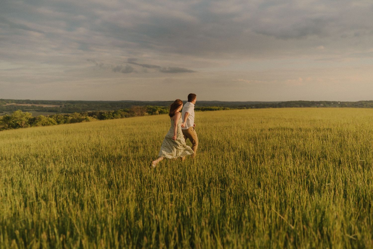 Two people walking through a golden wheat field at sunset with dramatic cloudy sky.