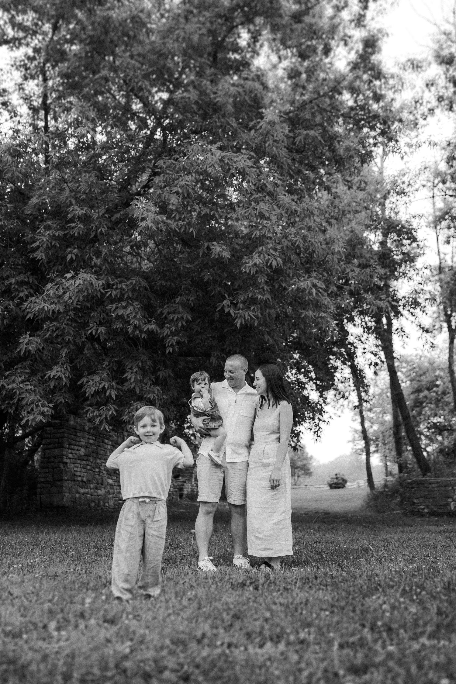 A tender outdoor family moment captured in black and white beneath lush trees on a grassy lawn.