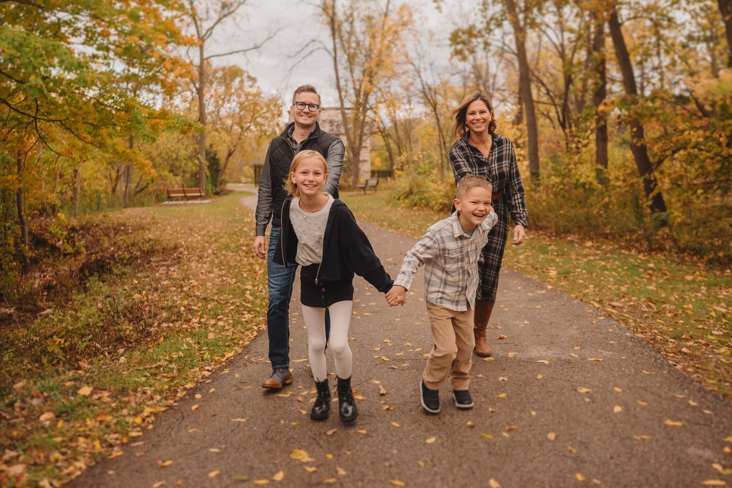 A family walks together on a leaf-covered path through autumn woods with golden foliage overhead.