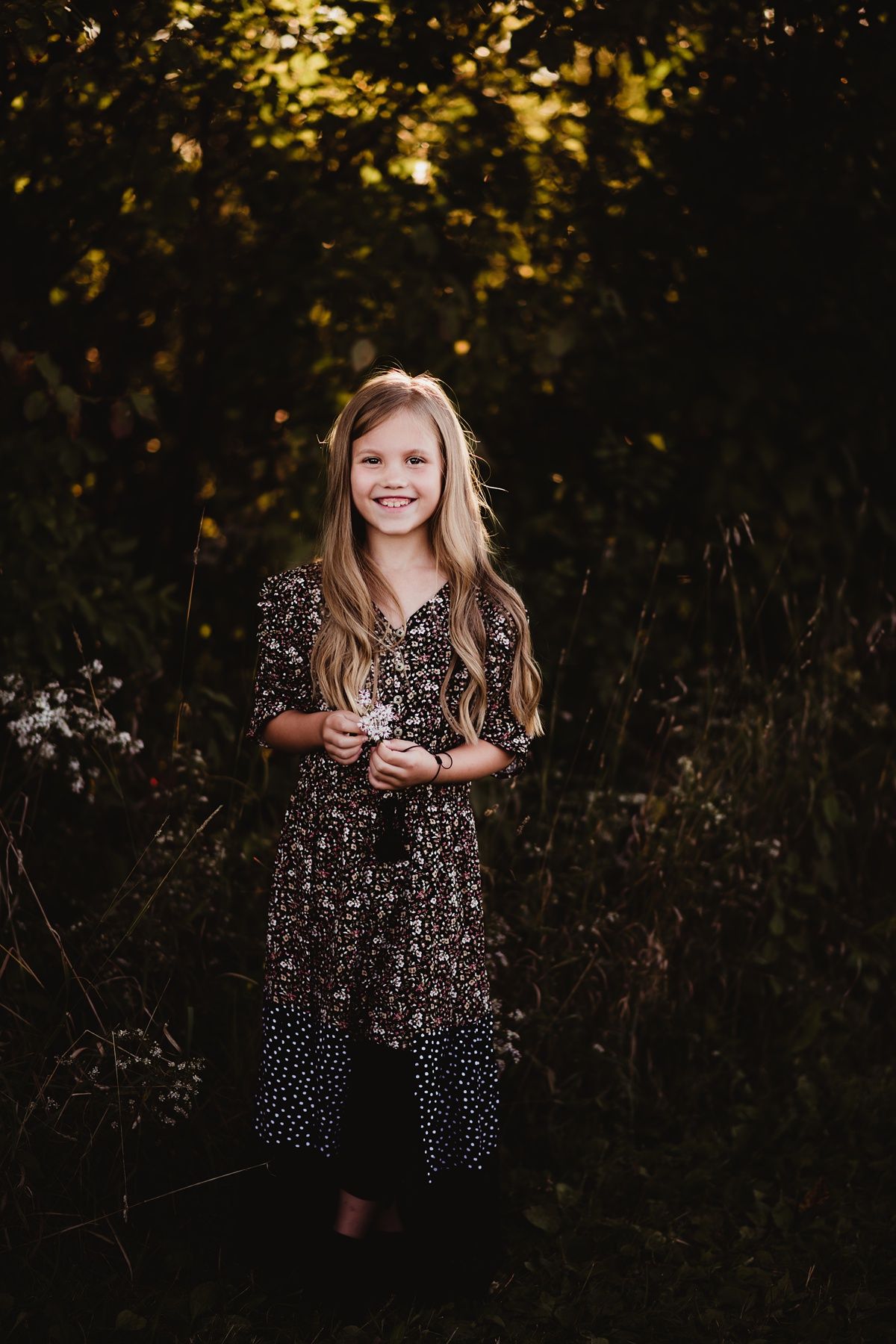 Child in floral dress standing in natural outdoor setting at sunset.