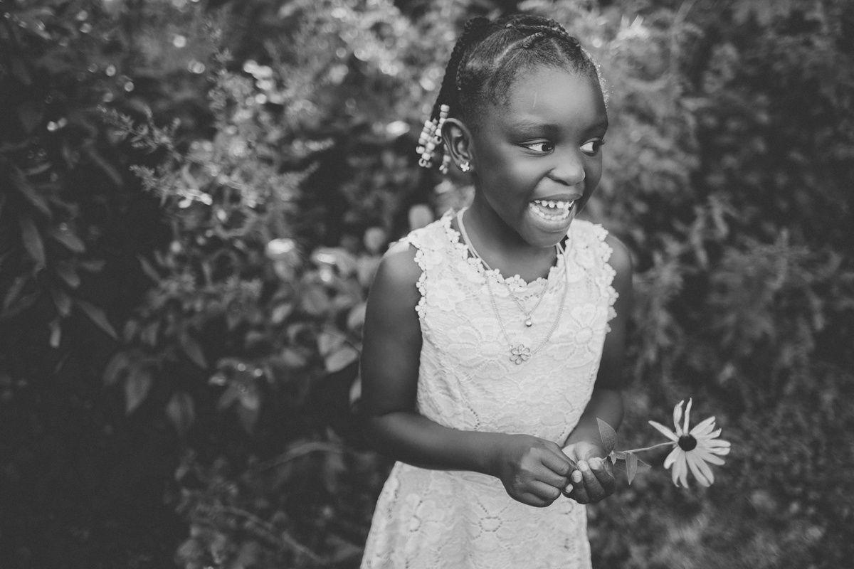 A black and white portrait showing joy while holding a flower against natural foliage.
