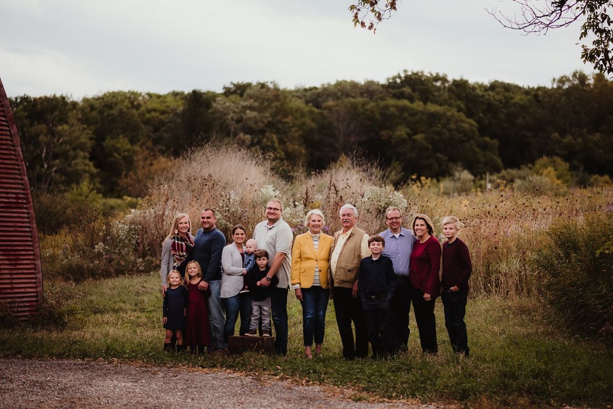 A multi-generational family gathering poses together outdoors in front of autumn foliage.