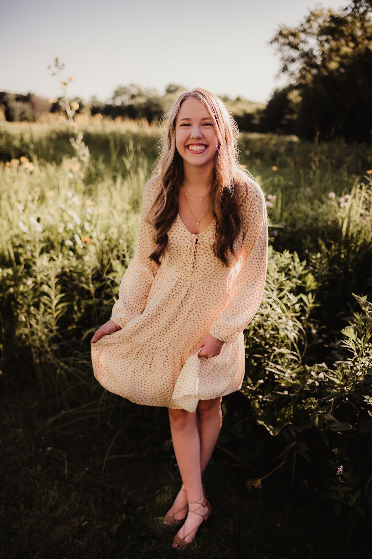 A dreamy golden hour portrait of a person in a flowy cream polka dot dress standing in tall wildflowers.