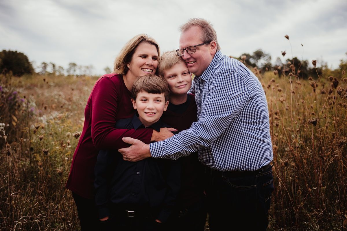 A loving family of four embraces while taking a portrait in a field of tall autumn grass.