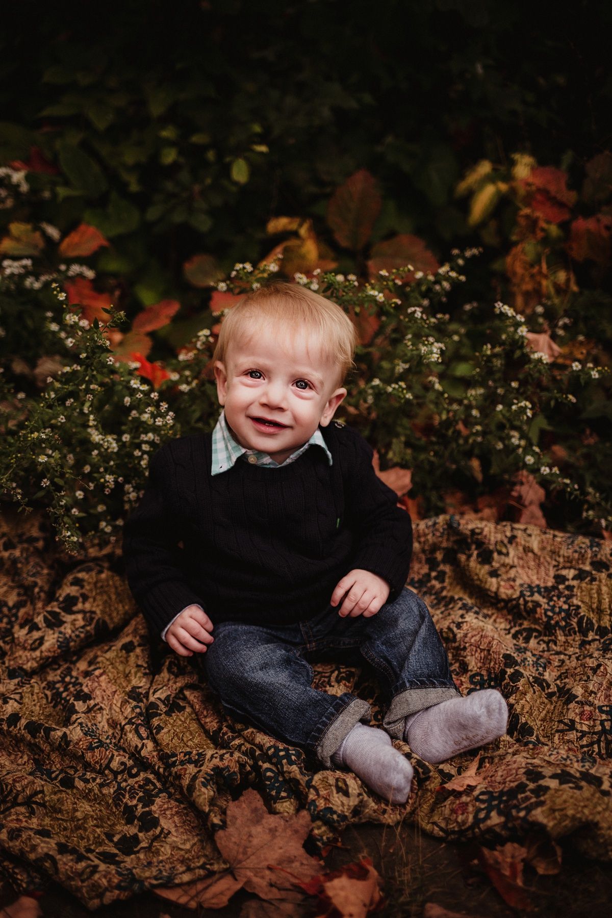 Young child in black sweater sits smiling among autumn leaves and flowers outdoors.
