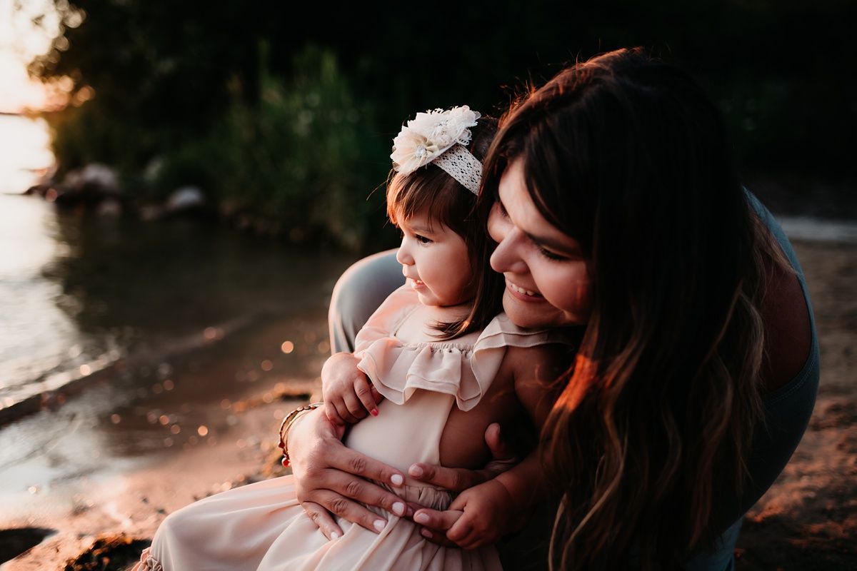 Intimate waterside portrait of parent and child in matching white dresses at golden hour.