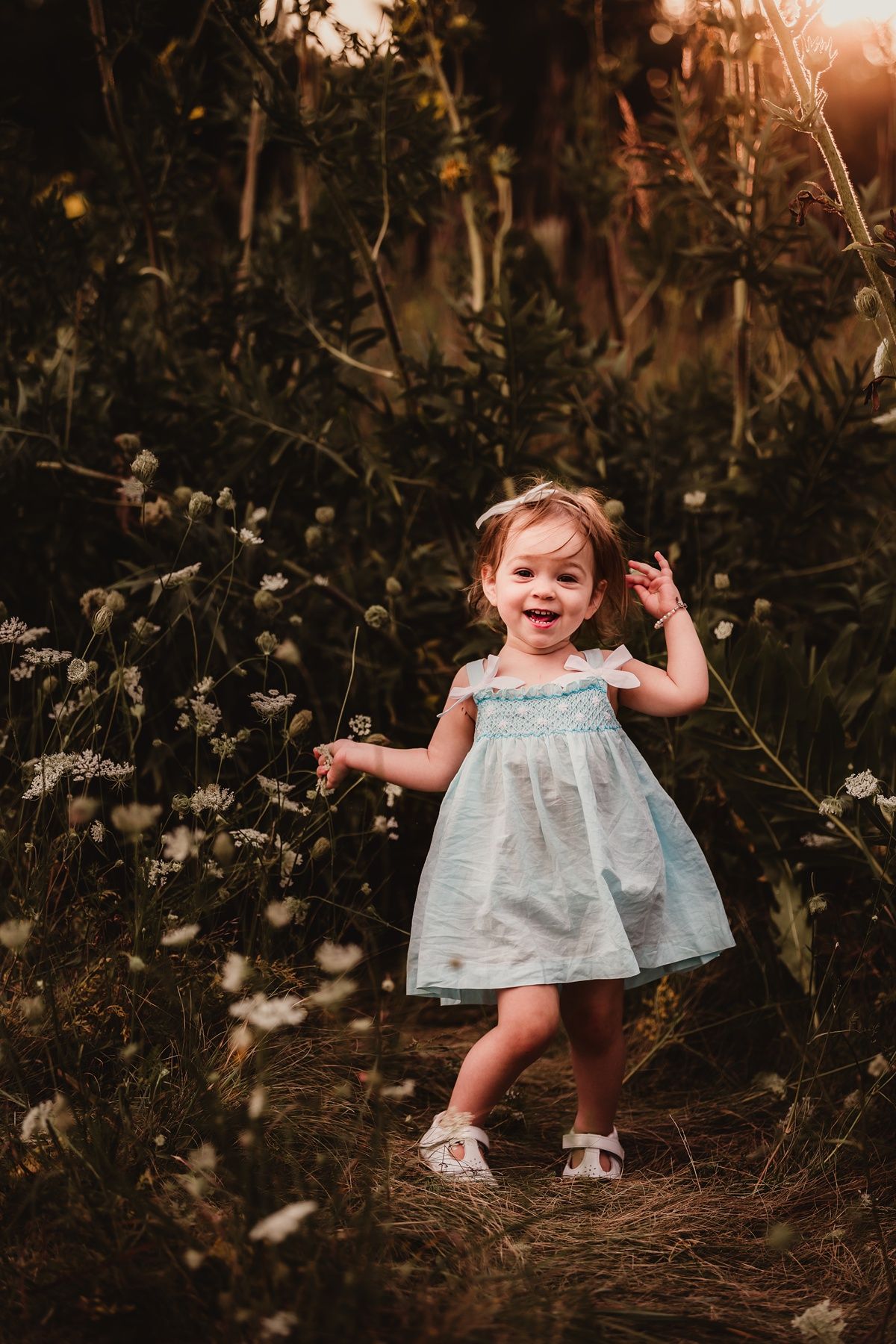 Joyful toddler in light blue dress plays among flowering bushes at sunset.
